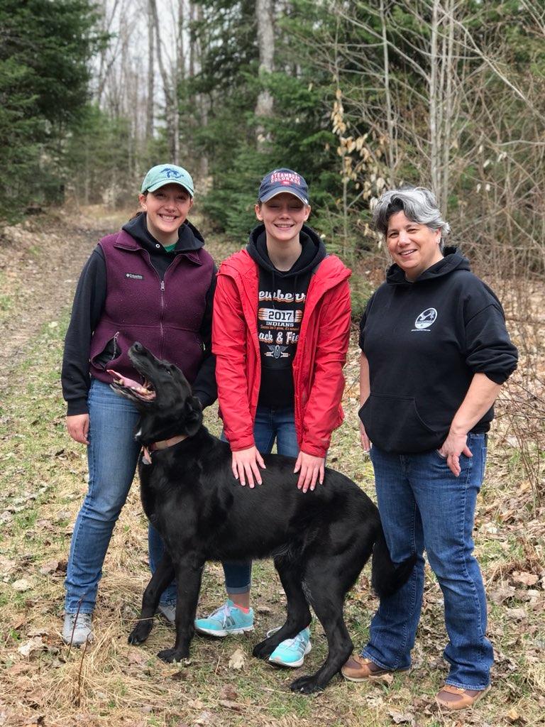 three women, eryn, her sister Carlee, and her mother Caryn wearing spring jackets and jeans, standing behind a large black lab named jack on a hiking trail. 