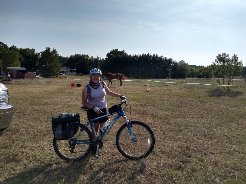 white woman, eryn, wearing a purple tank top and black bike shorts, standing next to her mountain bike with two full rear panniers. standing in a field with a grazing horse in the background. 