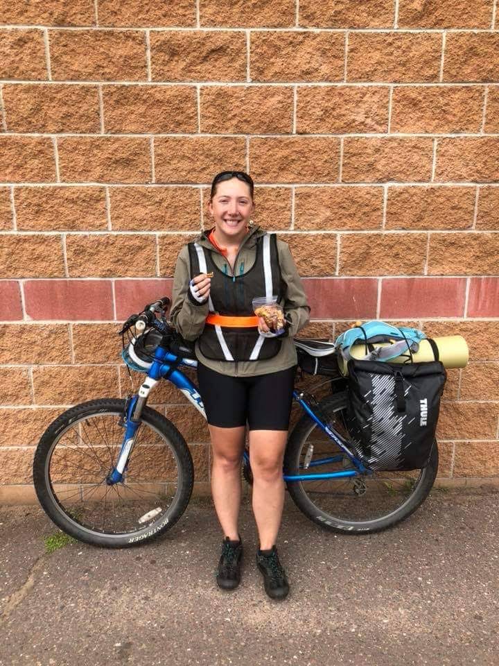 white woman, Eryn, standing in front of her mountain bike with full rear panniers, holding pretzels and smiling at the camera. bike and eryn standing in front of a brick wall.