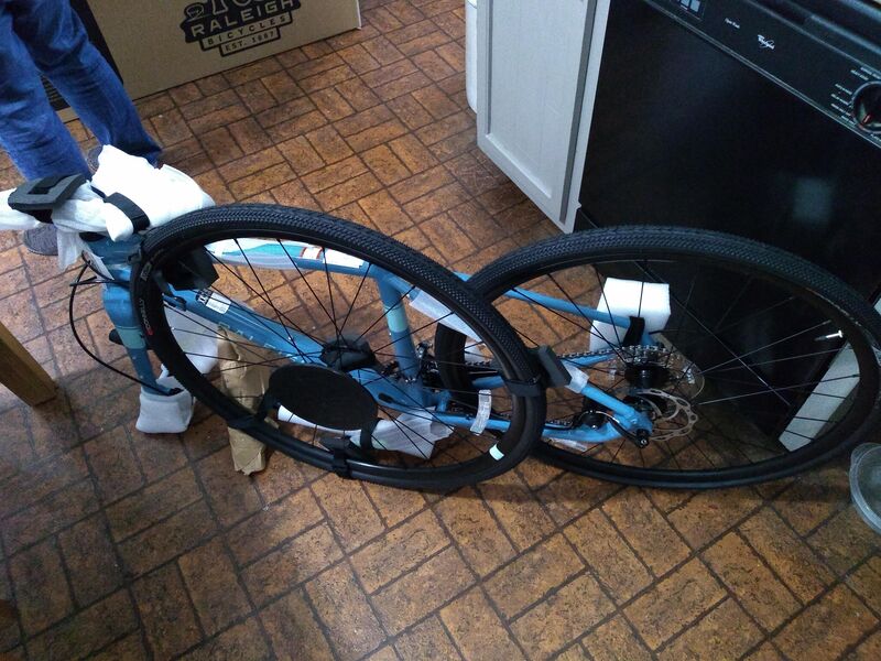 A deconstructed blue gravel bike taken out of a box. The front tire is not attached to the fork, it has styrofoam around the disc break and the handlebar is attached to the fork with plastic and more Styrofoam. The bike is sitting on the brown linoleum kitchen floor. 