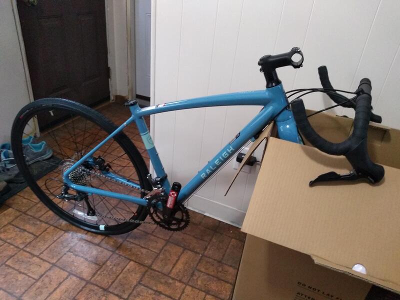 The blue gravel bike is part way put together. the bike is standing up, with the front fork over the side of the shipping box. the handlebars is resting on the top of the box with the forks. there is no seat or pedals on the bike yet. The bike and box is against a white wall, with the brown linoleum floor of the kitchen and shoes in the background.