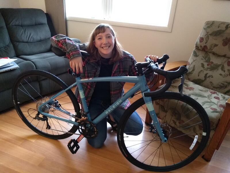 the blue gravel bike is fully constructed. Eryn, a white woman with shoulder length hair and bangs - wearing a flannel, black shirt, and jeans, is crouched behind the bike, with her left hand on the handlebar, and the right arm draped across the seat. in the background there are two couches and the floor is hardwood.