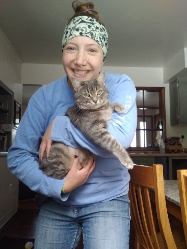 Eryn, wearing a blue long sleeve shirt, holding a grey striped cat, named Annie Oakley. 