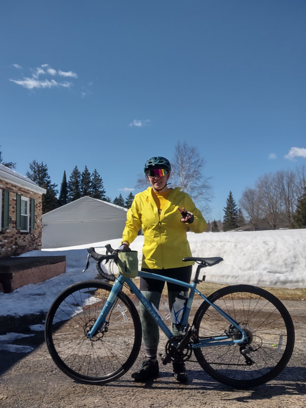 Me, eryn, wearing a bright yellow raincoat, reflective sunglasses, a green helmet, green merino wool leggings, purple striped socks, and black bike shoes standing behind the blue gravel bike. attached to the bike handlebars is a waterbottle. Eryn and the bike are standing infront of a 3ft snowbank. 