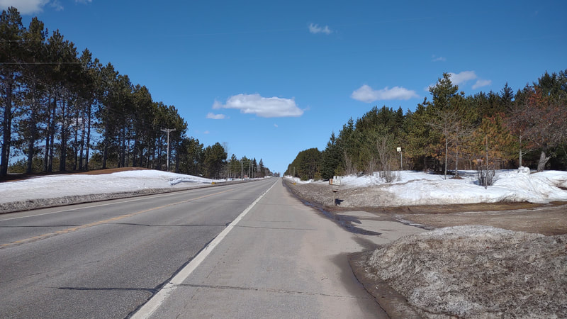 an empty 2 lane road with a wide shoulder. snowbanks on either side of the shoulders, with red pines lining the road. 