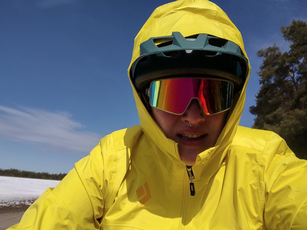 Eryn, with a grimace facial expression, wearing bright colored blender's wrap-around sunglasses, a green helmet, and a bright yellow raincoat with the hood up overtop of the helmet. sunny day, with blue skies and wispy clouds in the background. 