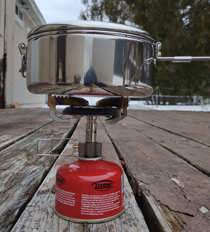 MSR minimalist silver cookstove connected to a Sterno ISO Butane/Propane fuel mix cannister. the stove is turned on, with a silver MSR pot with lid on top. this is all sitting on a wooden porch outside of a house. 