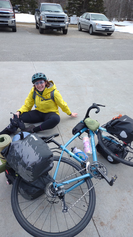 Eryn, smiling and sitting on cement with a yellow raincoat, black leggings, black shoes, holding black winter gloves, wearing a green helmet and blue glasses. In front of her is the blue gravel bike with full front and rear panniers, stem bags, and water bottles