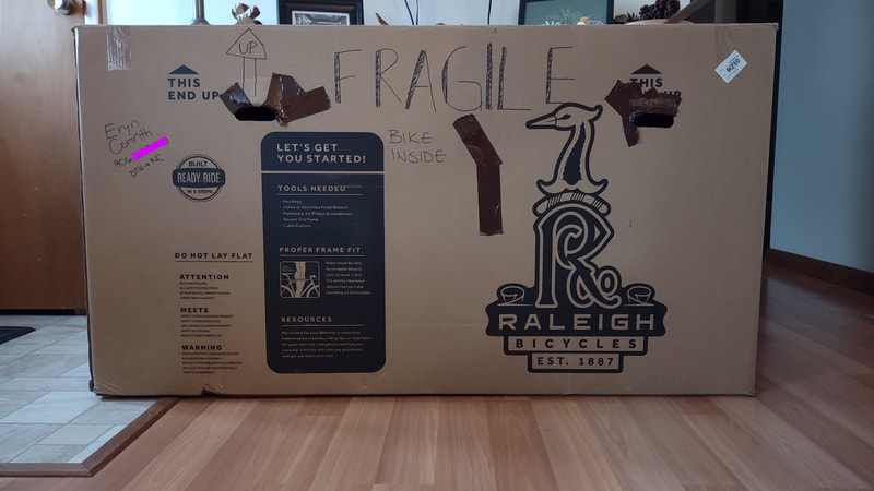 A large brown cardboard bike box that has Raleigh Bicycles logo and instructions. handwriting on the box that says "Fragile, bike inside" and "Eryn Corinth, redacted phone number, and DTW to RIC."