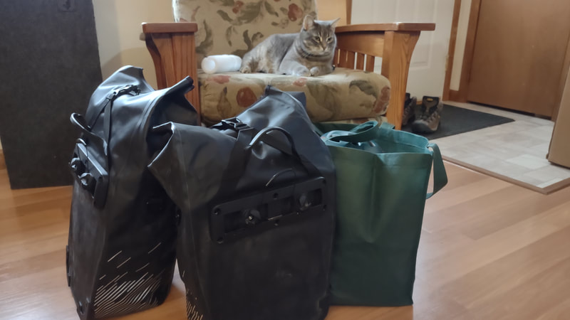 Two full black rear panniers and a full green reusable bag Infront of a lay-z-boy chair with a cat (Annie) lounging on it.