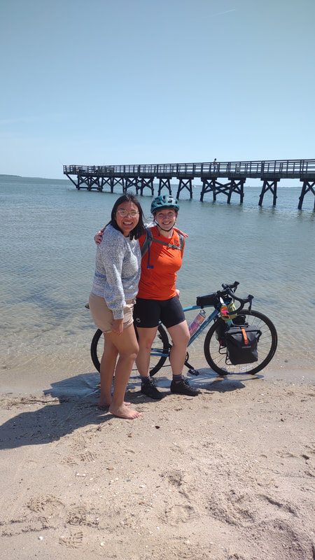 Eryn, wearing a bright orange shirt, black cycling chamois, purple striped socks, black shoes, and a green helmet standing next to her friend Clarisa who is wearing a blue and white long sleeve shirt, tan shorts, and is barefoot. both girls looking at camera and smiling. Clarisa and Eryn are standing infront of Eryn's fully loaded bike Infront of the ocean. The bike tires are in the ocean.