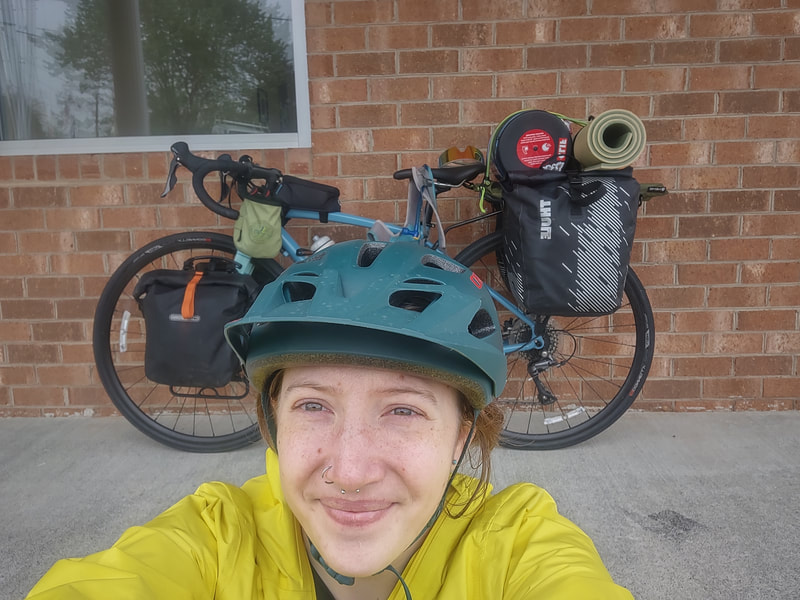 Eryn, taking a selfie, smiling at the camera with helmet & yellow raincoat on, behind her is her fully loaded bike resting on a brick wall.