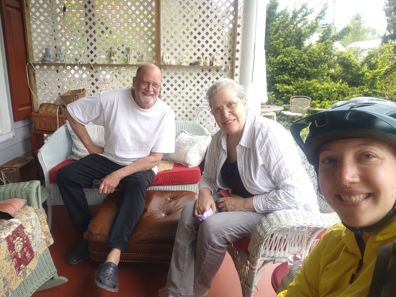 Eryn taking a selfie with two new friends, Wayne and Joan, who both are in their 70s. both are wearing white shirts and dark pants, Eryn is wearing her helmet and yellow rainjacket. all three are sitting on chairs on a porch.