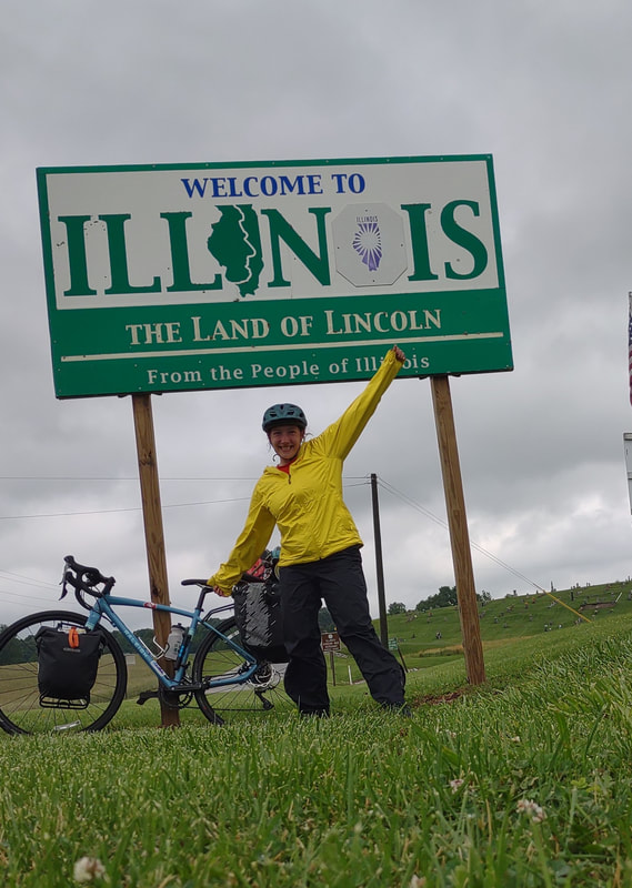 road sign that says "Welcome to Illinois. Land of Lincoln. From the people of Illinois." bike is leaning on the sign. Eryn is infront of the sign with full rain gear on in a slanted pose with arms spread out