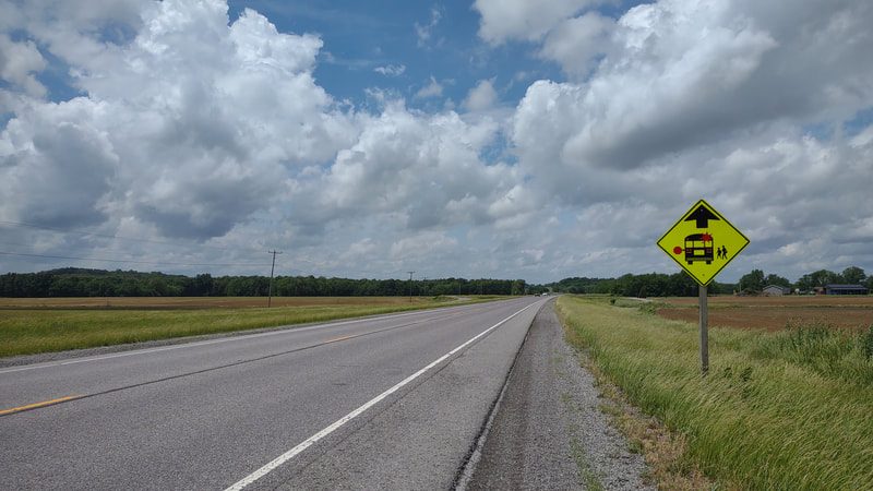 two lane highway, gravel shoulder, grass ditch and then farm field with small corn growing. large voluminous clouds on horizon. school bus road sign on right hand side.