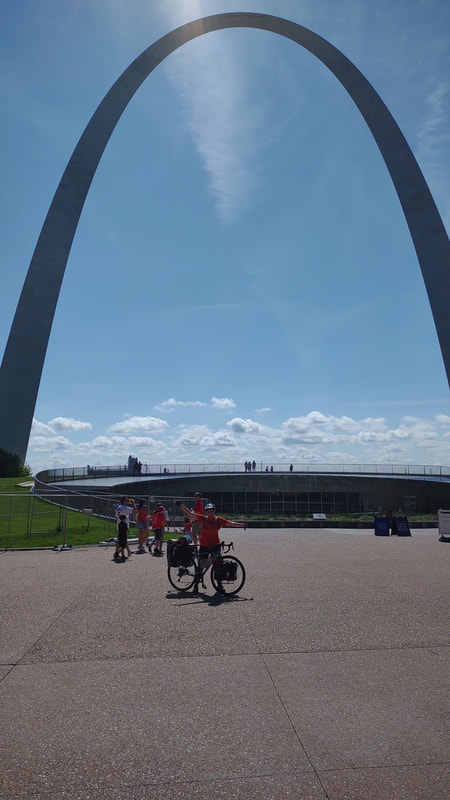 St. Louis arch in the background, Eryn and fully loaded bike in the foreground. the bike is learning on Eryn and her arms are spread out reaching for the sky.