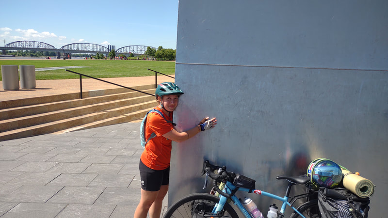 Eryn hugging a corner of the St. Louis arch, bike is leaning on the arch as well. 