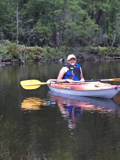 Eryn, a white woman, wearing a black/red lifejacket sitting in a red kayak on a calm river. 