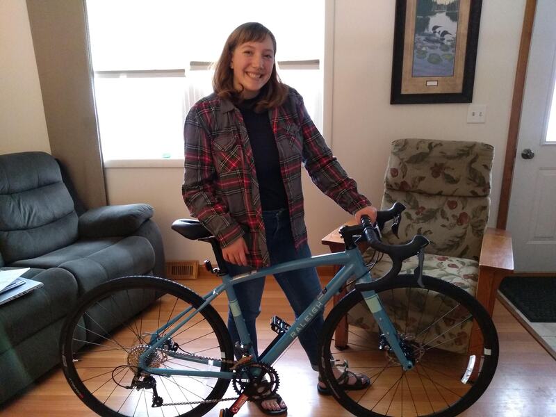 eryn standing wearing a red/grey flannel and blue jeans, holding onto a new blue gravel bike in her living room. 