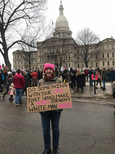 Eryn wearing a pussycat standing in front of a crowd with protest signs and the Michigan capitol building at a 2018 women's march in Lansing Michigan. 