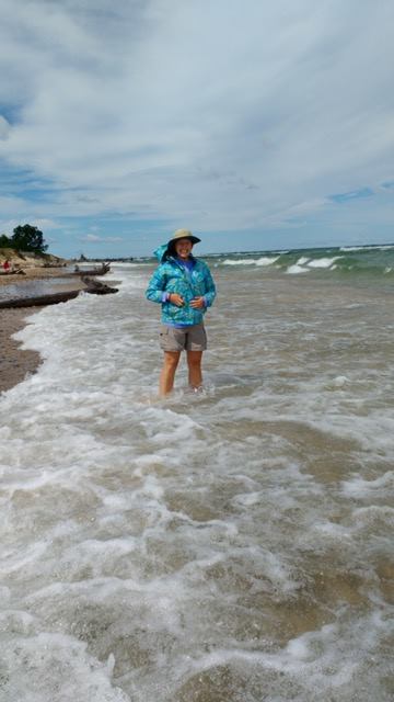 A woman standing in a swell of Lake Superior, in a blue raincoat, bucket hat, and tan shorts.